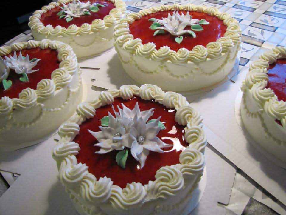 Monta valkeaa kermalla koristeltua kakkua, joiden päällä on punaista kiillettä ja massasta tehtyjä valkeita kukkia