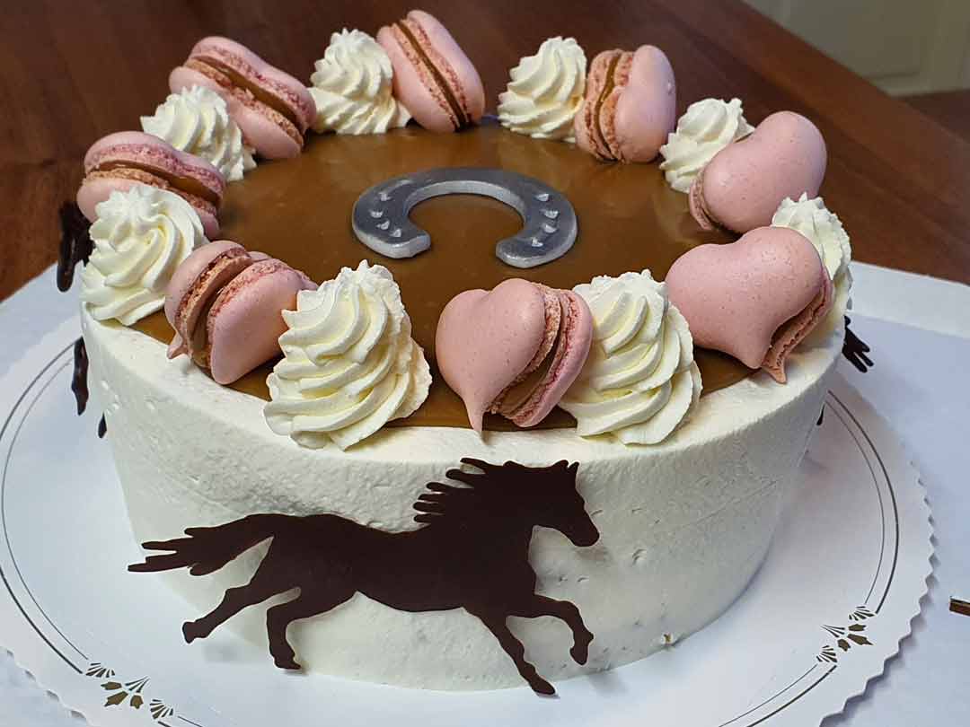 Valkea kakku, joka on koristeltu hevosaiheisilla koristeilla, kermakierteillä ja sydämen muotoisilla macaron-leivoksilla
