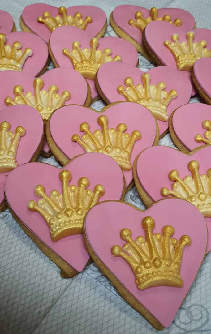 Sydämen muotoisia keksejä, jotka on koristeltu vaaleanpunaisella kuorrutteella ja kultaisilla kruunukuvioilla